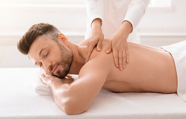 Obraz premium Unrecognizable woman therapist rubbing joyful man shoulders at modern spa salon, side view. Handsome bearded middle aged man relaxing while getting healing back massage