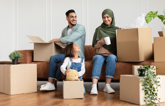 House Move And Relocation Concept. Portrait Of Cheerful Family Housing, Parent And Happy Smiling Little Girl Packing Or Unpacking Cardboard Boxes Together In Livin Room, Sitting With Open Packages
