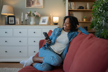 Obraz premium Smiling woman relaxing on sofa using smartphone laughing