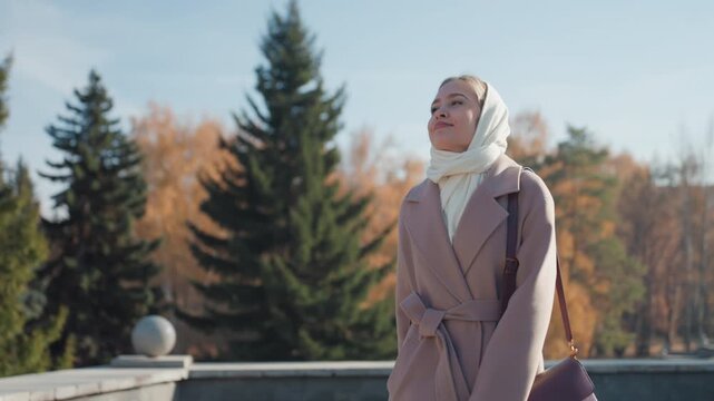 White woman in scarf on bridge, autumn park backdrop. Contemplative stroll as tourist and student, soft sunlight through pine trees, muted coat and stone balustrade, lamp posts, gentle smile, hands