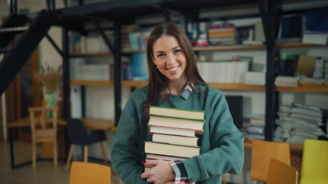 Student carries stack of books in library as she smiles while surrounded by shelves filled with more books