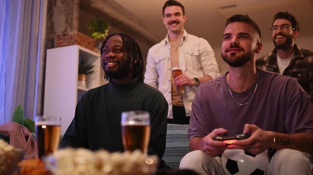 Cheerful multicultural friends playing soccer video game and drinking beer together at home in the evening