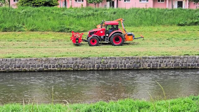 A professional red tractor equipped with a hydraulic arm flail mower performs seasonal grass cutting on a steep river canal slope.