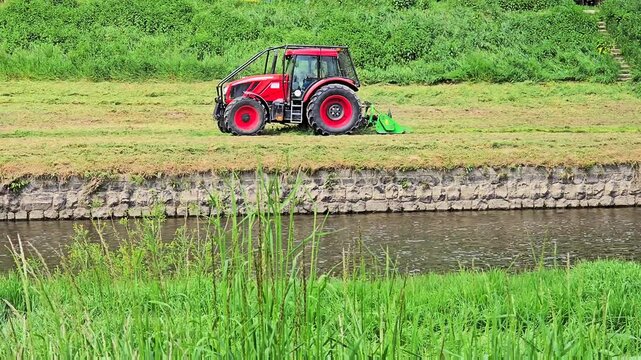 A professional red tractor equipped with a hydraulic arm flail mower performs seasonal grass cutting on a steep river canal slope.