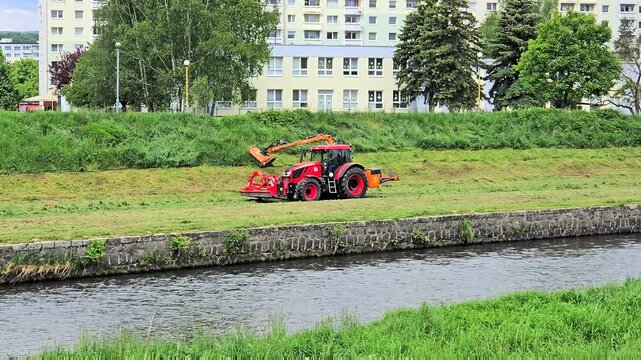 A professional red tractor equipped with a hydraulic arm flail mower performs seasonal grass cutting on a steep river canal slope.