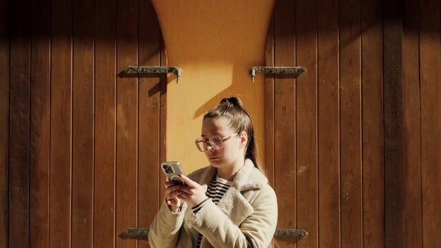 Young woman with glasses using smartphone near rustic wooden doors in golden sunlight. Travel lifestyle, navigation, urban explorer, old town.