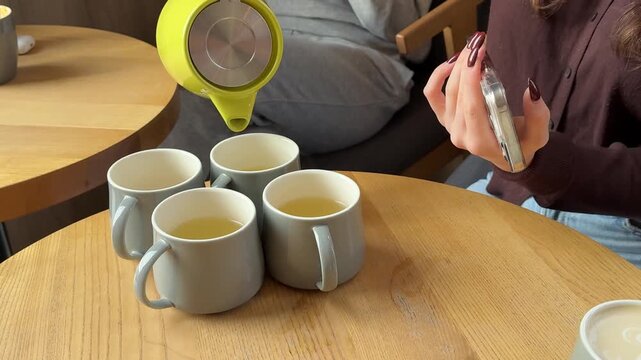 Hands of girl pour tea from green teapot into four cups on wooden table while recording on smartphone. Cozy cafe, beverage moment, and modern lifestyle concept.