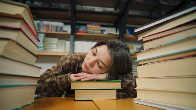 Person resting on books at a library during the day amidst stacks of books and study tables, surrounded by shelves of library materials