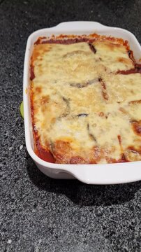 Vertical footage of a homemade freshly baked eggplant parmigiana, typical sicilian dish, over a dark marble kitchen countertop.