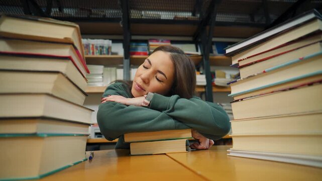 Student rests on desk in library surrounded by books during study session in the afternoon