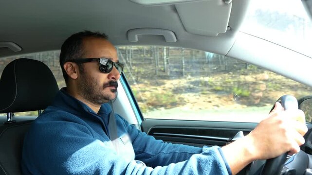 Portrait of a stressed man wearing sunglasses driving a car and feeling nervous or tense during a difficult traffic jam or long commute on a sunny summer day