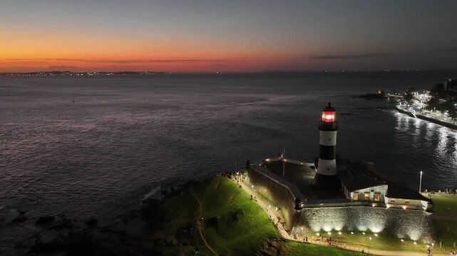  Aerial reveal of Farol da Barra lighthouse at dusk with red sky, glowing horizon, and ocean coastline