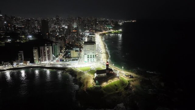erial reveal of Barra coastline and city skyline at night with illuminated buildings and Farol da Barra lighthouse in Salvador, Brazil