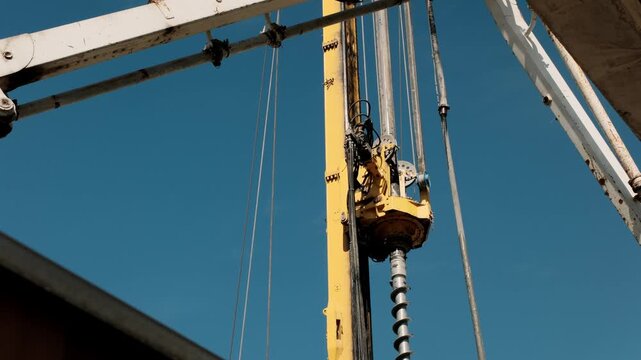 Construction drilling rig with auger and hydraulic rotary head. Low angle view of pile foundation drilling machine, cables and mast against clear blue sky. Industrial geotechnical drilling equipment.