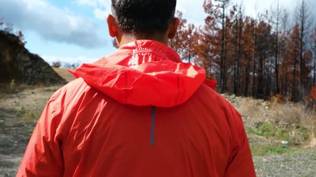 Rear view tracking shot of a man in an orange raincoat walking deep into a burnt and decimated forest area highlighting the ecological impact of wildfires