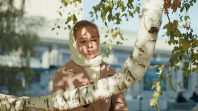 Young caucasian woman leaning on birch tree in city park with scarf, dappled sunlight and shadow patterns, distant station and architecture, thoughtful gaze as student waiting and remembering, cool