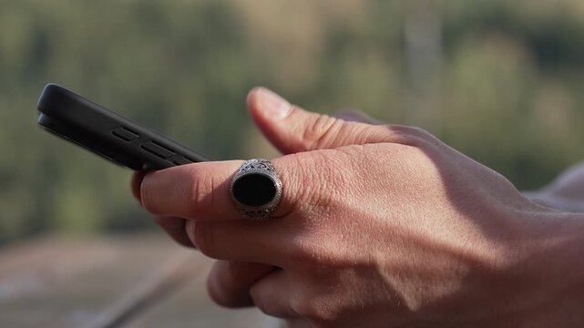 Close-up of a man's hands using a smartphone outdoors with a focus on a stylish silver ring with a black stone, natural background.