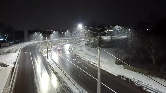 Winter, snow is falling. Car drive of the slippery road. Lanterns on poles illuminate the road in the evening time
