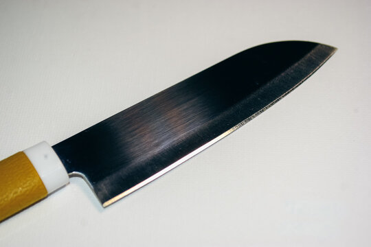 Sharp blade gleams under bright light. Yellow handle contrasts with sleek metal. Minimalist background highlights knife's craftsmanship. Ideal for cooking, cutting, or kitchen design themes