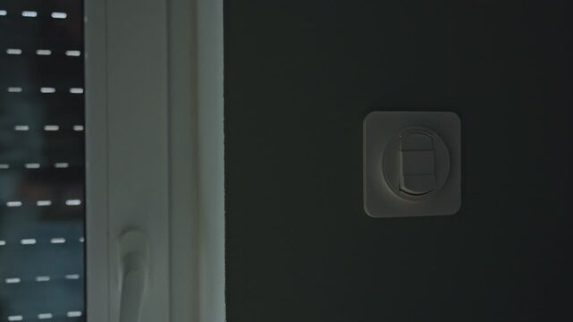 Woman pressing wall switch while exterior roller shutter goes up on window. Concept of light control, privacy and residential security