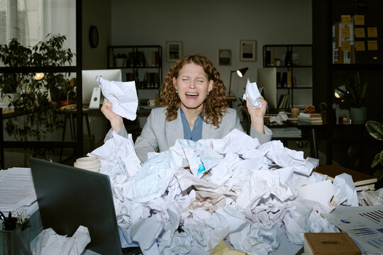 Caucasian young woman sitting at cluttered desk holding crumpled papers, appearing stressed while surrounded by large pile of documents in modern business office environment