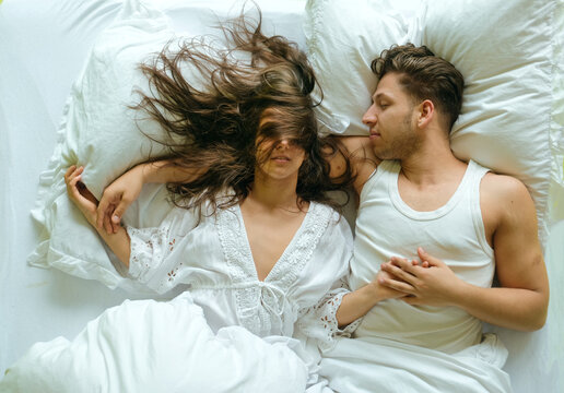 Young couple lying in bed holding hands woman hair flowing across face soft natural light creates intimate authentic moment of connection tenderness and quiet morning atmosphere