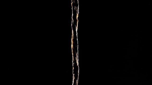A thin stream of golden liquid flows downward against a stark black background creating a striking visual contrast