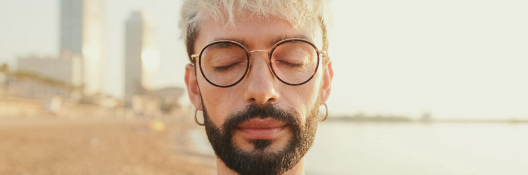Close-up of a young guy with glasses standing on the beach with his eyes closed. Panoramic