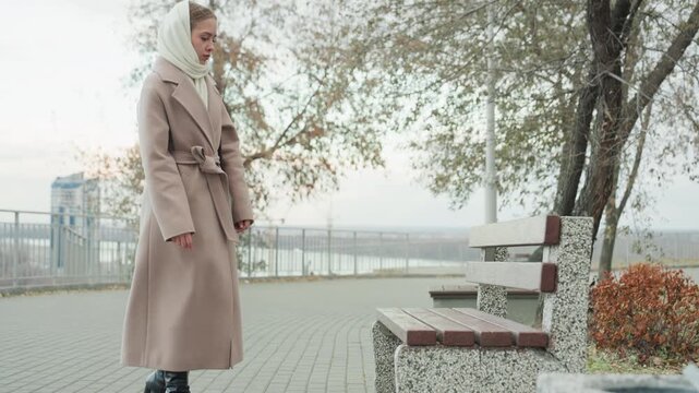 Full body woman walking bench side, commuter pausing to sit and reflect, coat cinched at waist and scarf tucked, leaves on pavement and waterfront railing, calm overcast sky and soft seasonal light,