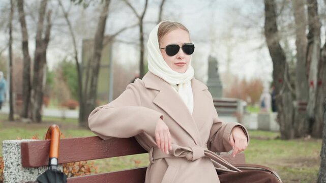 Elegant caucasian woman on bench wearing beige coat and white scarf, adjusting sunglasses while vintage umbrella rests beside her in quiet autumn park poised model vibe, serene contemplative mood