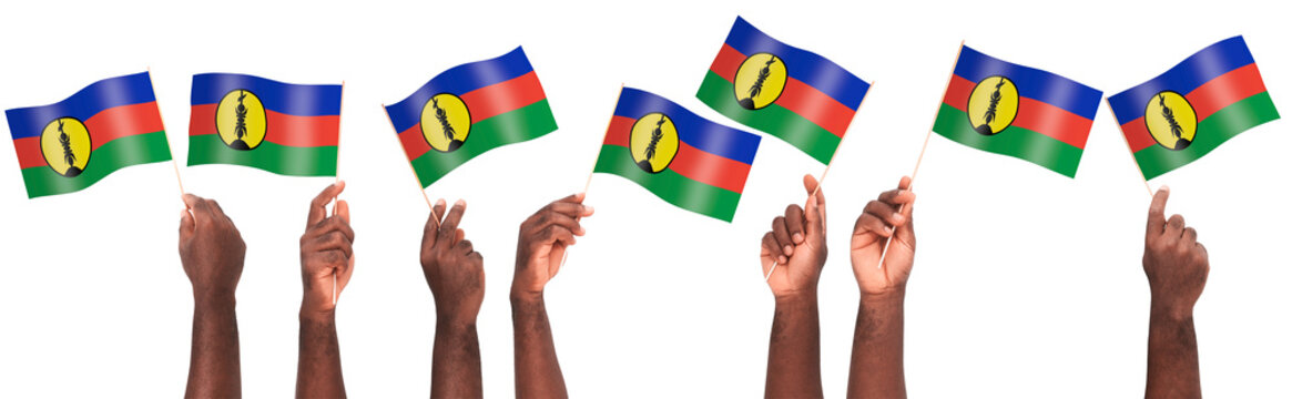 Multiple hands hold the New Caledonian flag aloft against a white backdrop. The vibrant blue, red, and green stripes with the yellow sun emblem symbolize Kanak identity and national solidarity.