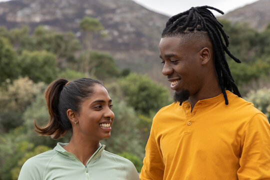 Diverse couple smiling and facing each other while standing on trail in hills wearing activewear