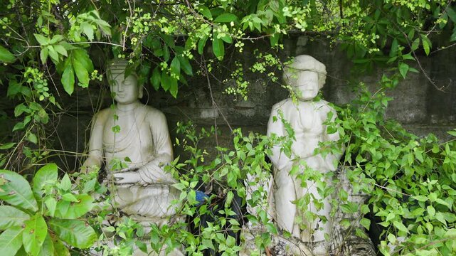 Two stone statues, one seated and one standing, are partially obscured by foliage in a garden in Ngu Hanh Son, Da Nang, Vietnam. The statues are weathered and covered in moss.
