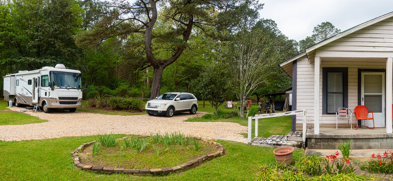 A camper van is parked in a rustic setting beside an old wooden house. The scene is framed by vibrant garden flowers and a stone porch.
