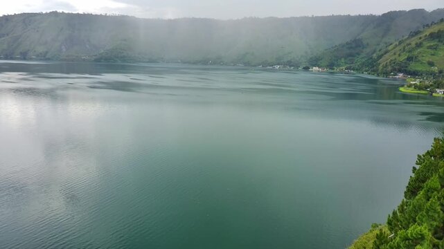 4K drone footage of Lake Toba showing calm turquoise water, surrounded by green hills, pine trees, and light mist, creating a peaceful and cinematic natural landscape.