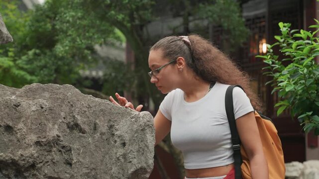 Multiracial woman inspecting stone up close, researcher like focus as she touches lichen and studies surface texture beside courtyard trees, glasses and notebook imply educational fieldwork and quiet