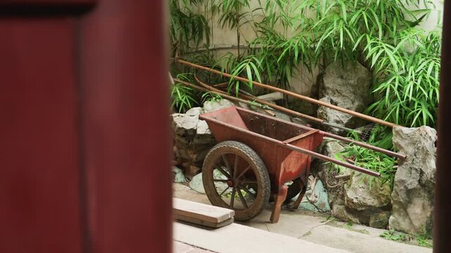Rustic wheelbarrow beside stone wall bamboo, red doorframe in foreground, wood planks on ground, piled rocks and gardening tools leaning, soft daylight filtering through foliage, weathered paint