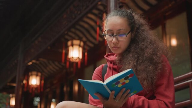Young woman reading under lanterns in covered pavilion with warm lighting and closeup focus evening study aesthetic with glasses, curled hair, thoughtful expression and creative notetaking