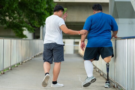 Caregiver assisting and taking care of disabled man with prosthetic leg outdoors. Supportive friend caring for male amputee with artificial limb near public stairs in urban.