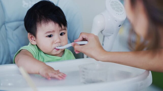 Happy Asian baby boy in high chair smiling at mother while being spoon-fed. Close-up view with natural light in a home setting.