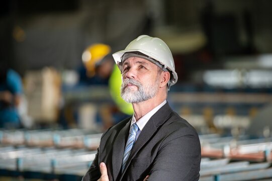 older owner engineer with beard mustache on face standing smile with his arms crossed at construction factory site. Boss manager in suit show good thumb at factory industry site.