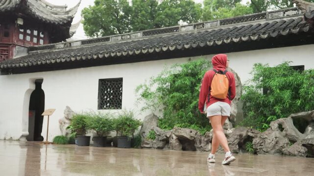 Asian woman tourist walking temple garden red hoodie orange backpack moving slowly across wet courtyard, passing circular moon gate, rockeries and potted bonsai mood contemplative serene, soft
