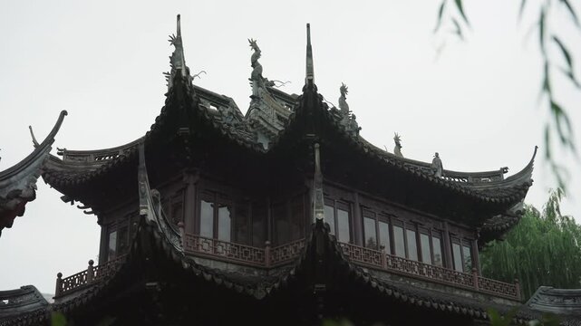 Rainy pagoda rooftop under overcast sky, ornate curved eaves and tiered tiles framed by willow branches, dark timber carving and decorative brackets, moody soft light casting deep shadows, historic