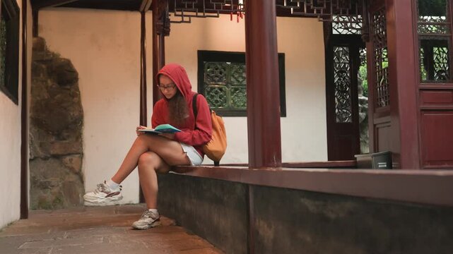 Asian woman reading book in pavilion, backpacker student journaling on stone ledge, warm evening light, wooden columns and carved screens, textured plaster walls, casual hoodie and sneakers, focused