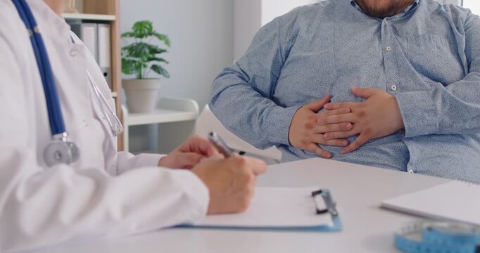 Doctor listens as a patient with obesity shows pain in the abdomen during a clinic consultation. Obesity patient reports stomach pain to physician sitting at desk in medical office.