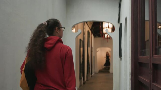 Young woman in red hoodie walking through arched gallery corridor carrying yellow tote, long ponytail, warm lantern light casting soft shadows, moody interior, candid back view as she explores