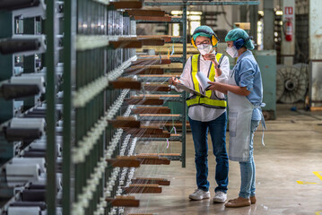 Textile inspection factory teamwork female supervisor in safety vest checking and discussing yarn production quality with woman worker in textile mill holding thread spool and clipboard. © BESTIMAGE