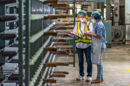 Textile inspection factory teamwork female supervisor in safety vest checking and discussing yarn production quality with woman worker in textile mill holding thread spool and clipboard.