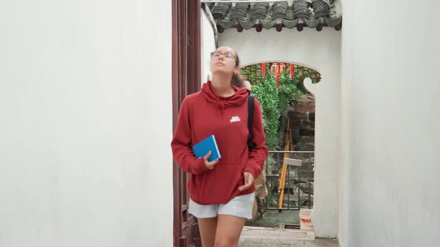 Young latina student with notebook walking through historic courtyard doorway, wearing red hoodie and holding blue journal while glancing at architectural details and hanging lanterns, candid study