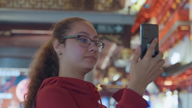 Woman with phone under lanterns, captivated traveler in red hoodie and glasses explores vibrant indoor market, scrolling map app and photographing traditional architecture warm bokeh lights and ornate